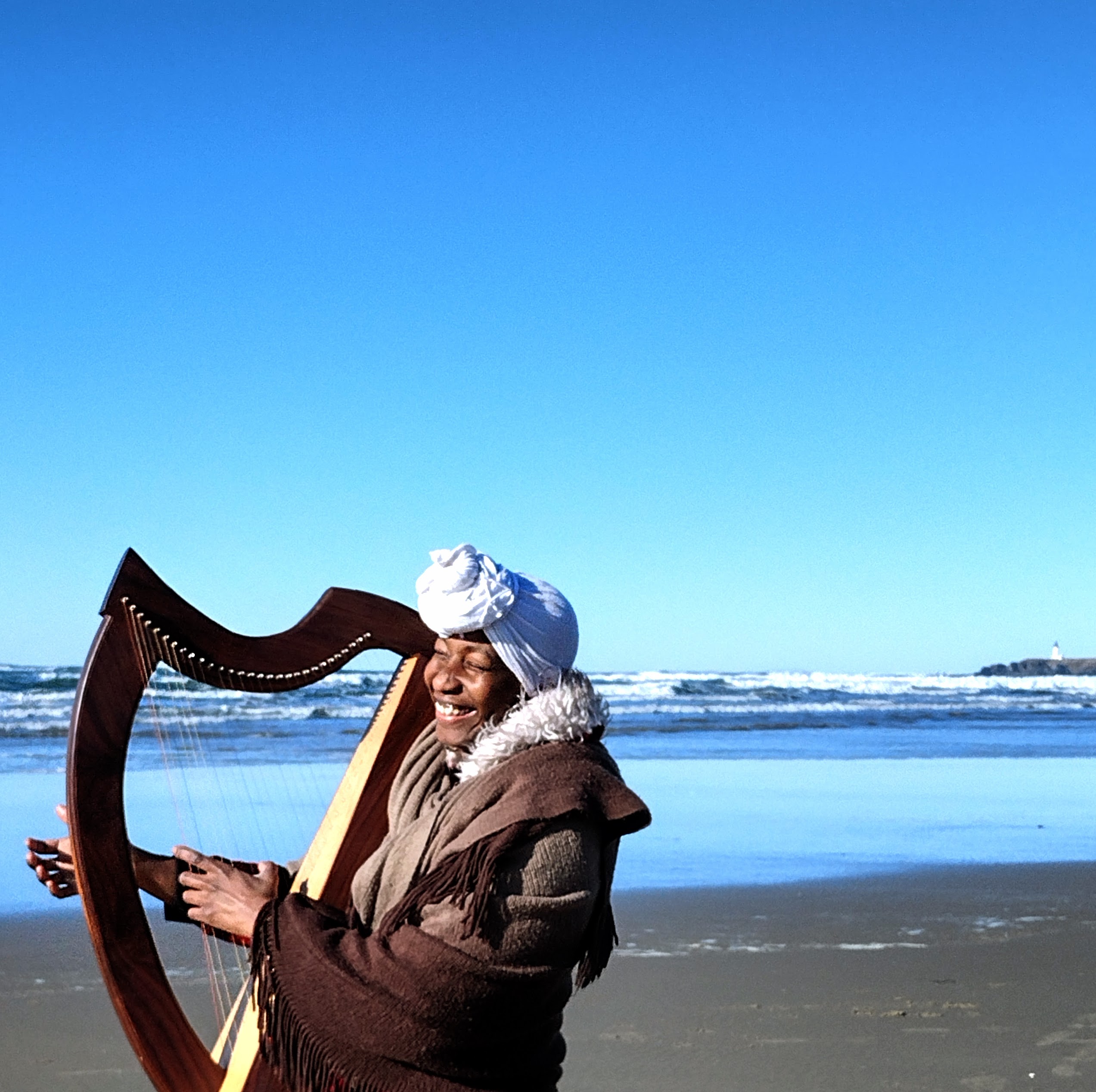 Maya Thornton playing a lever harp on the beach, lighthouse in the distance